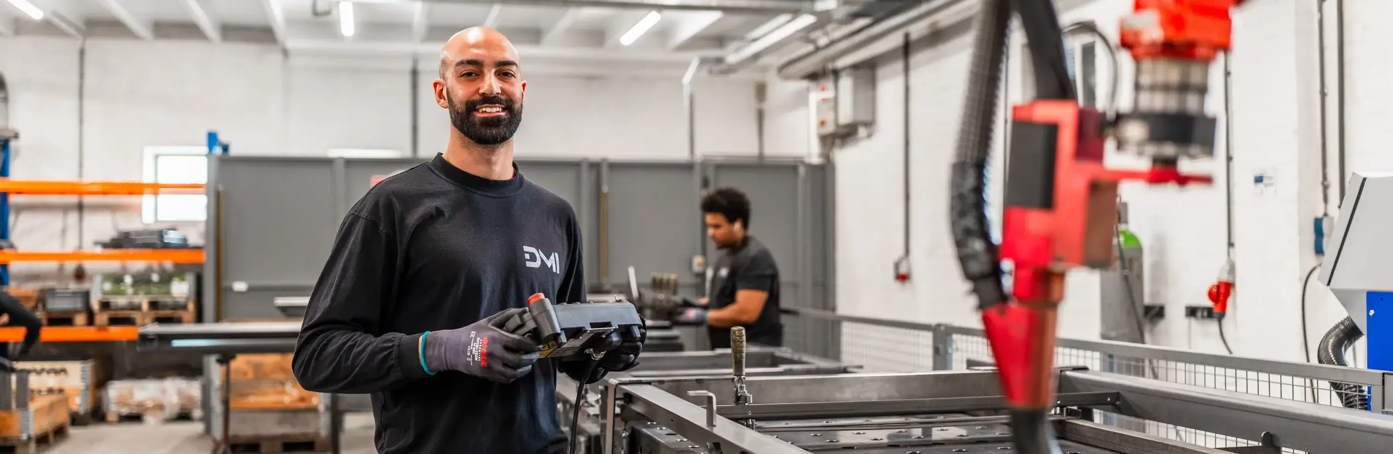 Glimlachende man in zwarte werkkleding houdt een bedieningsunit vast naast een rode lasrobot in een industriële werkhal.