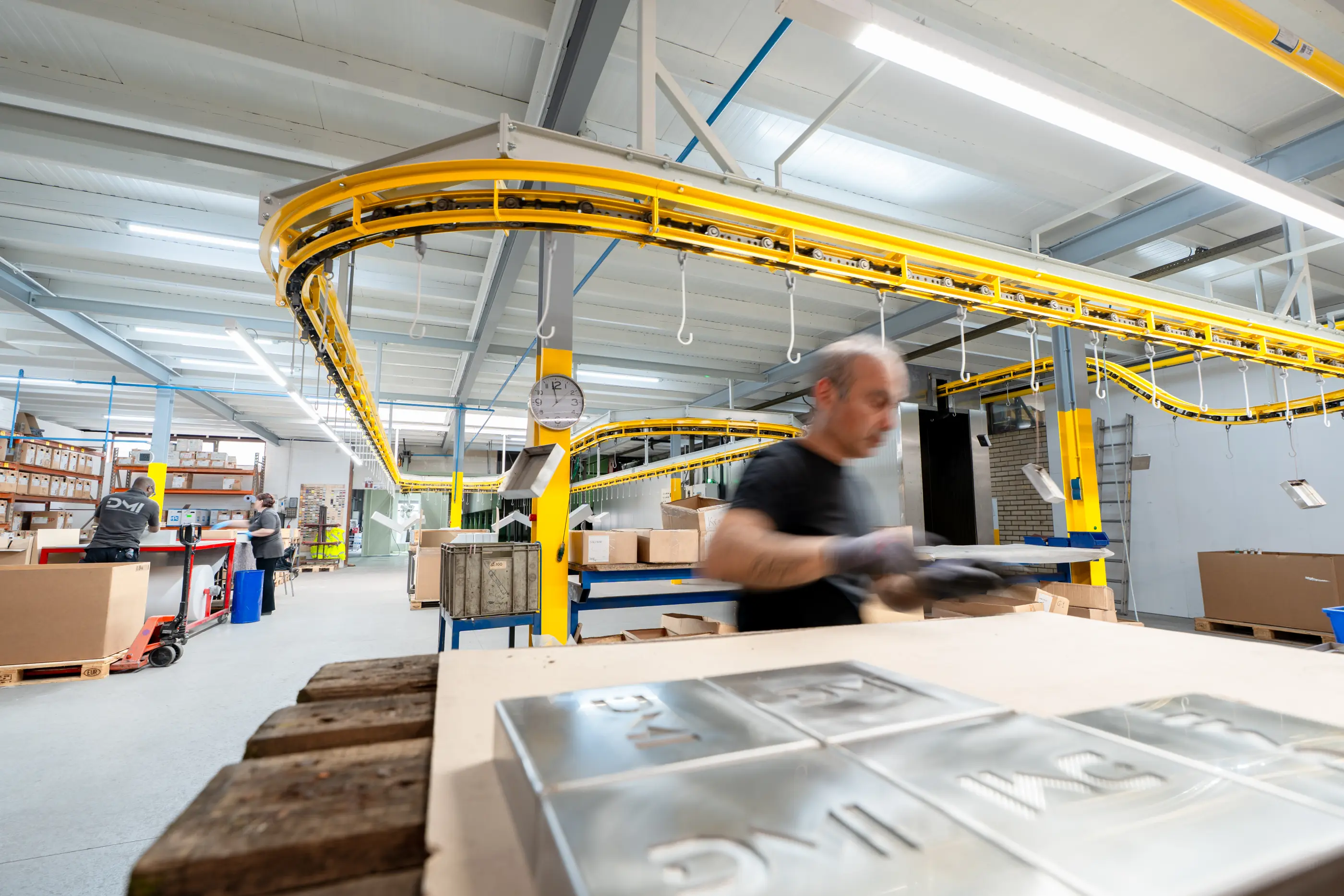 Medewerker met handschoenen werkt aan metalen platen in een lichte fabriekshal met gele hangende transportbaan en dozen.