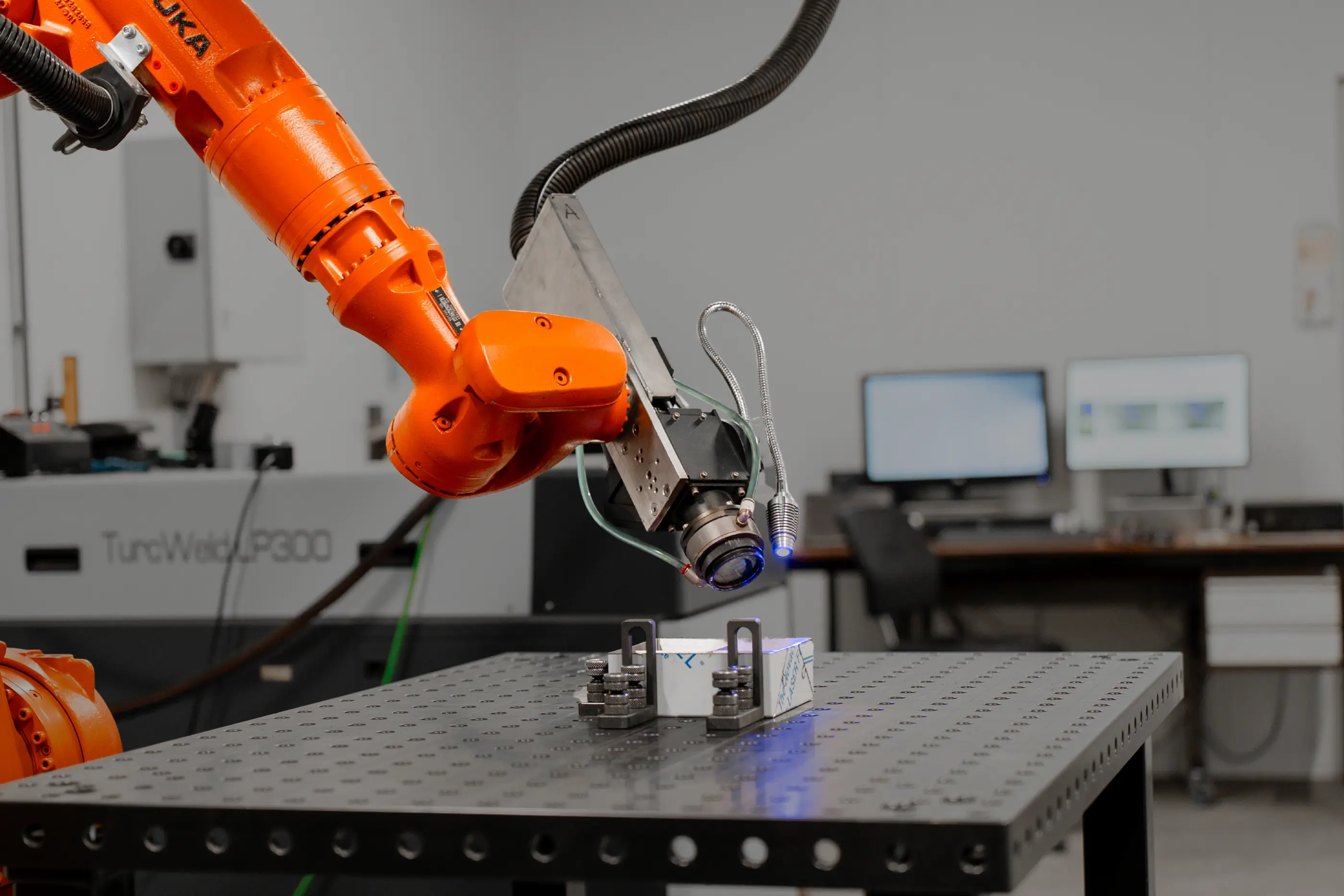 Oranje KUKA-robotarm met lasoptiek boven een geperforeerde werktafel waarop een werkstuk is vastgeklemd, in een werkplaats.