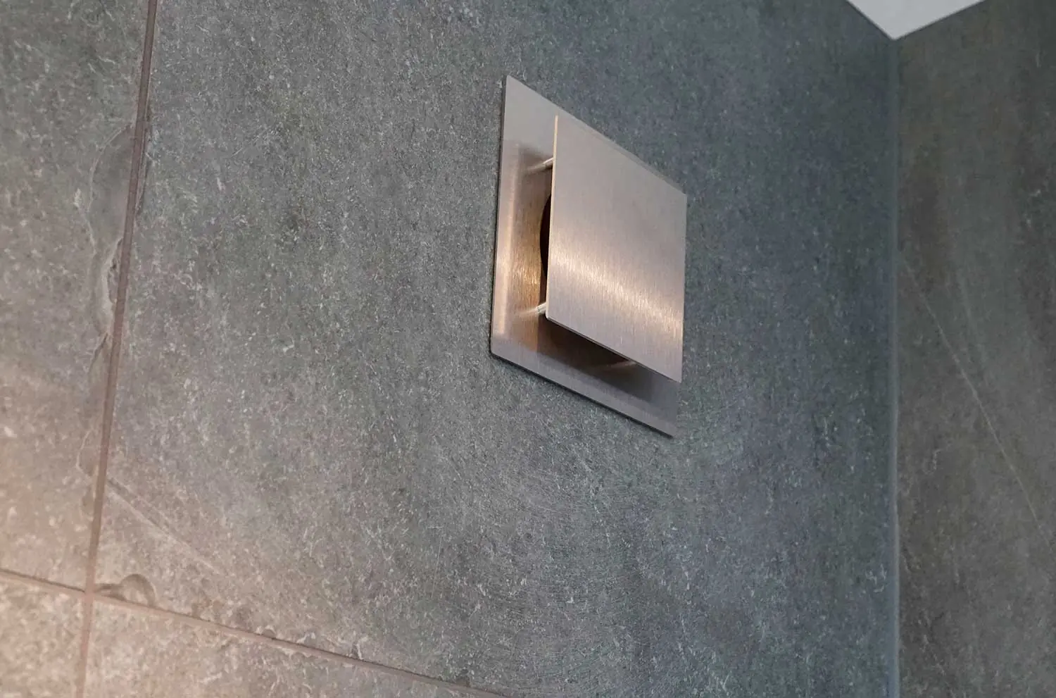 Vierkant geborsteld metalen ventilatierooster met open klep in een wand van grijze, gestructureerde natuursteentegels.