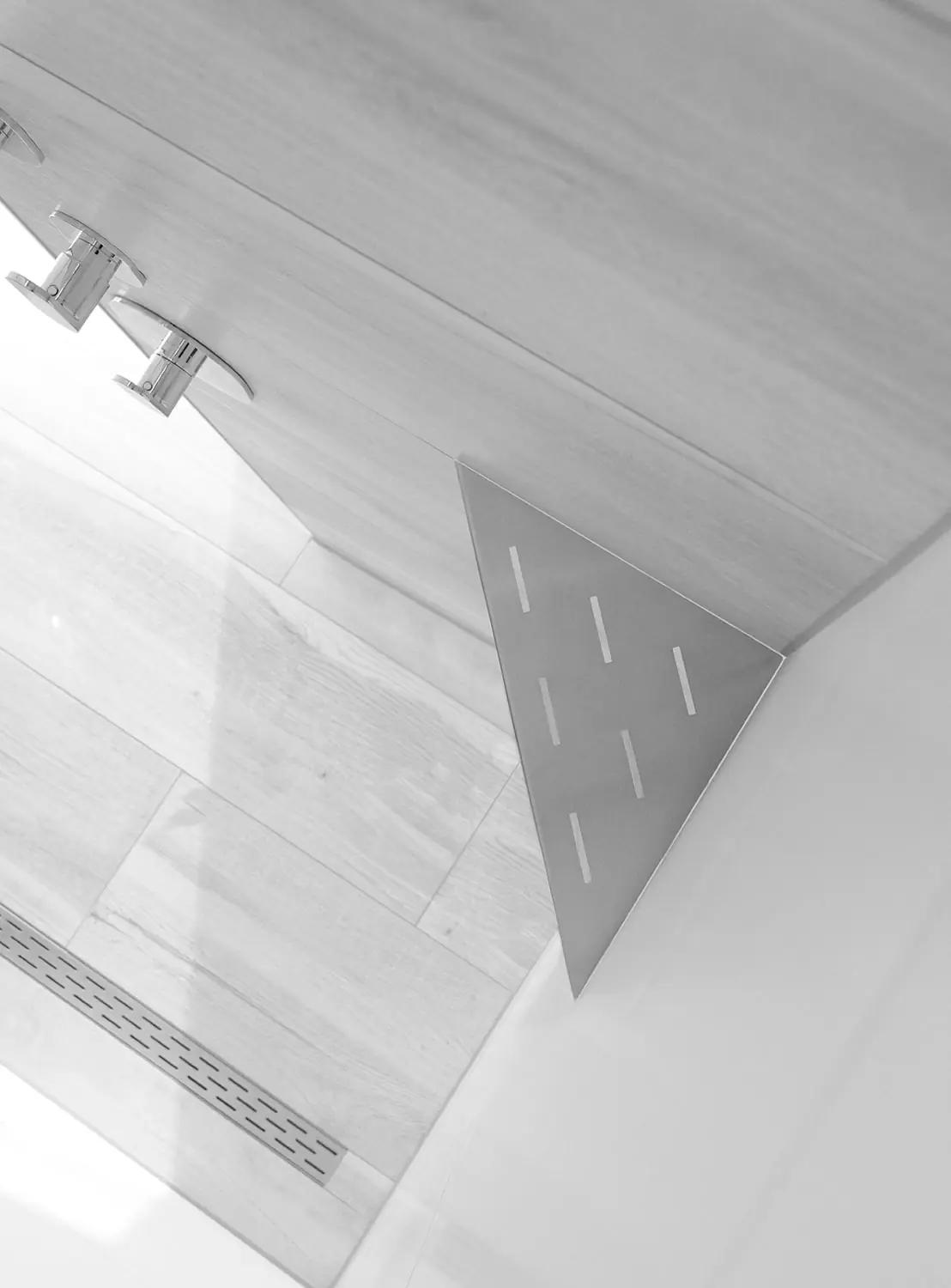 Zwart-witbeeld van een douche met houtlook tegels, driehoekige roestvrijstalen hoekplank, chromen kranen en een lineaire afvoergoot.