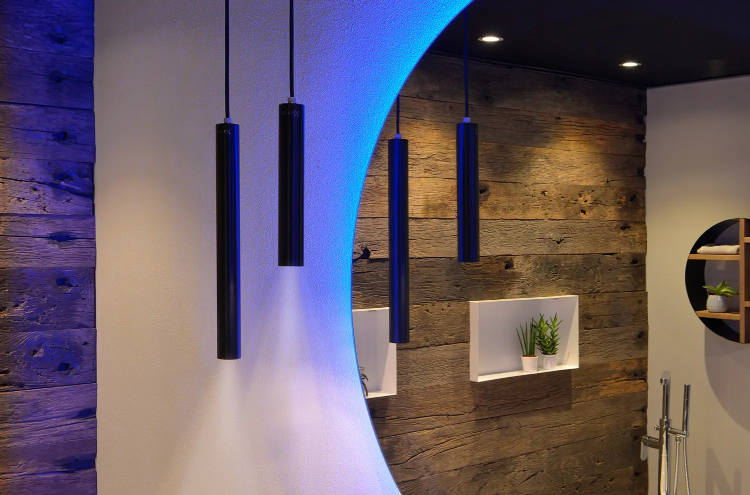 Zwarte cilindervormige hanglampen voor een ronde spiegel met blauwe ledverlichting en een houten wand met witte nissen en planten.