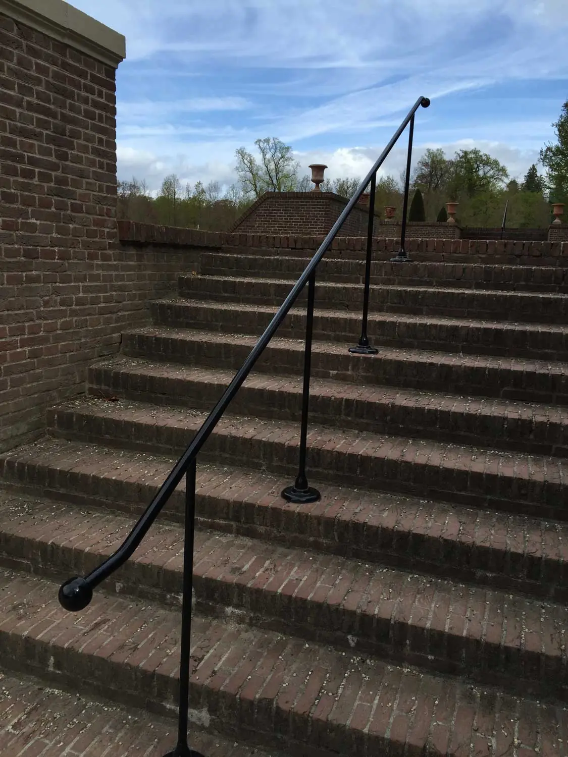 Zwarte metalen trapleuning langs een bakstenen buitentrap met terracotta vazen op de muur en een blauwe lucht erboven.