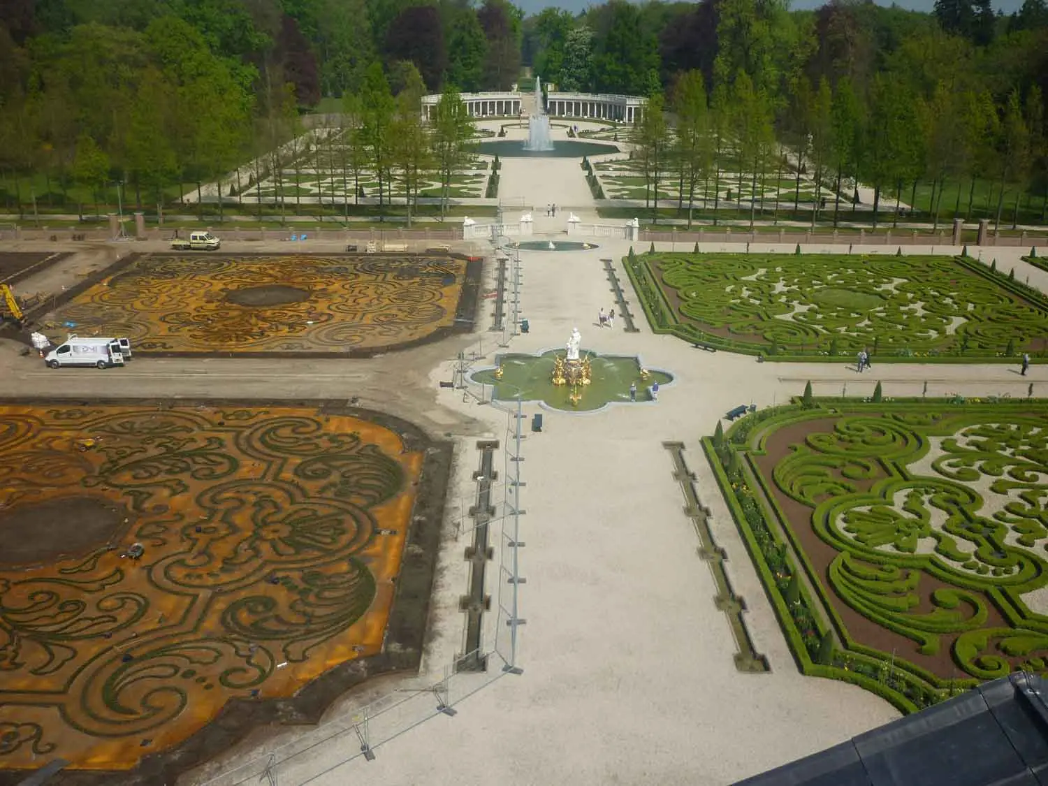 Bovenaanzicht van een symmetrische barokke tuin met sierlijke heggenpatronen, fonteinen en een kolonnade; links wordt nog gewerkt.