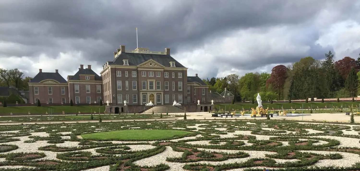 Bakstenen paleis met trappen en symmetrische siertuin met kiezelpaden onder een dreigende, donkere wolkenlucht.